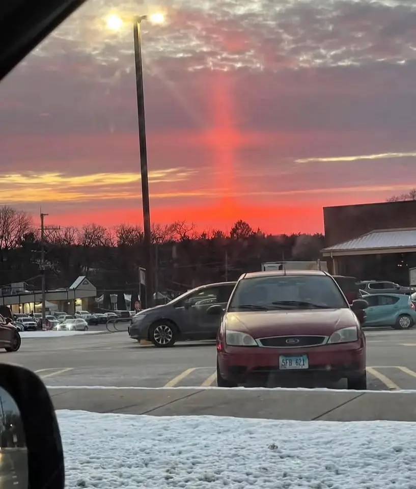 A photograph taken from inside a car shows a parking lot at dusk with snow on the ground. A striking red-orange light beam…