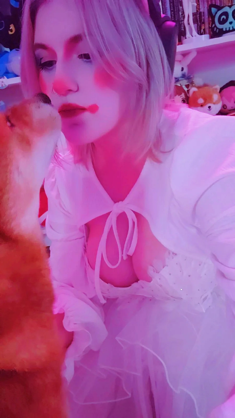 A person with short blonde hair and a nose ring, wearing a white ruffled top and skirt, is bathed in pink light while a…