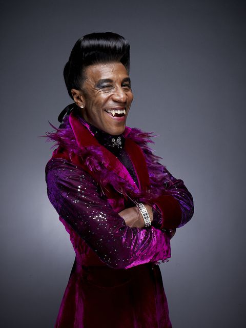 a man with a big smile and arms crossed, wearing a sparkly purple velvet outfit with feather trim and a diamond bracelet…