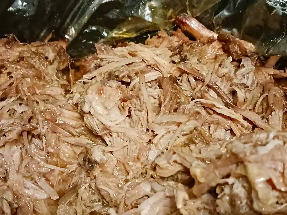 A close-up photo shows shredded, moist-looking pulled pork inside a dark, reflective slow cooker pot. The meat is fibrous…
