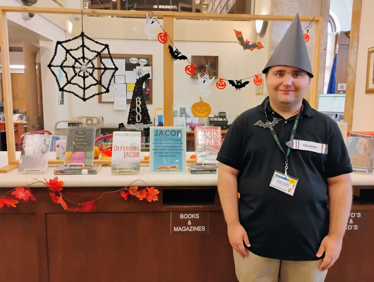 A man named Jacob, wearing a grey witch hat and a name tag that says 'Jacob', stands in front of a library counter decorated…