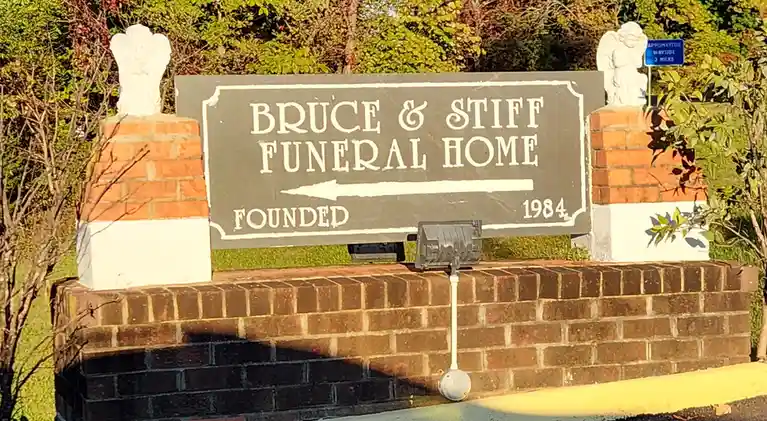 A dark gray sign for 'Bruce & Stiff Funeral Home' with an arrow pointing left and 'Founded 1984' is mounted on a brick wall…