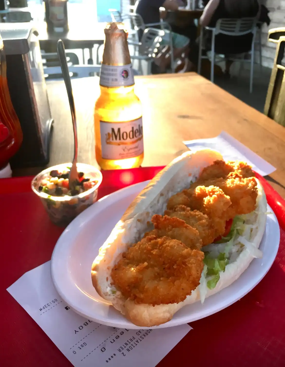 A close-up photo of a fried shrimp sandwich on a white plate, served with a side of salsa and a Modelo beer bottle on a red…