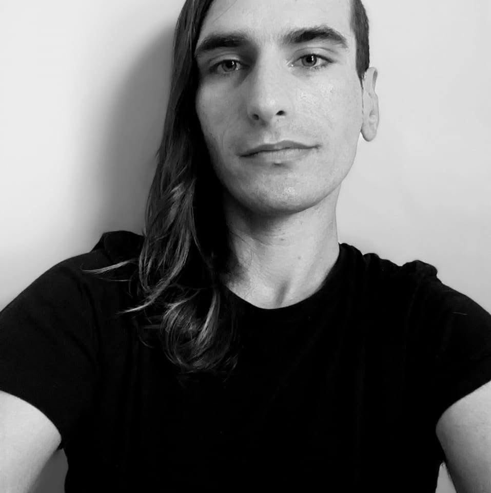 A black-and-white selfie of a man with long hair wearing a black t-shirt, looking directly at the camera with a neutral…