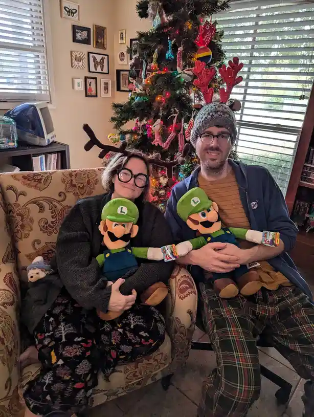 Two people sit on a patterned couch in front of a decorated Christmas tree, each holding an identical plush Luigi doll. The…