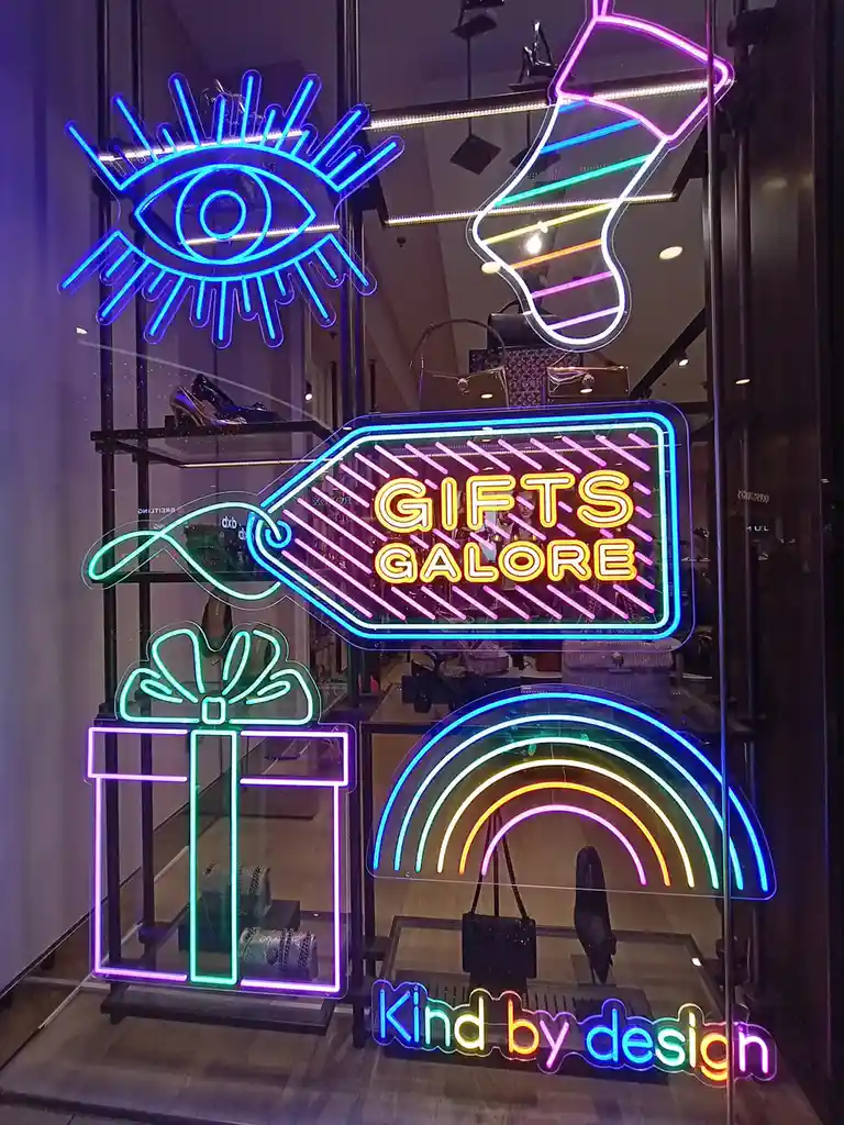 A vibrant storefront window display features colorful neon signs shaped like an all-seeing eye, a guitar, a gift box, a…