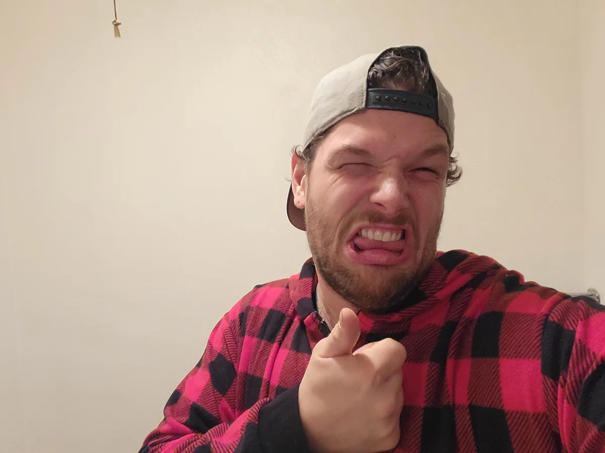 A man wearing a backwards grey baseball cap and a red-and-black plaid hoodie makes an exaggerated angry face with his eyes…