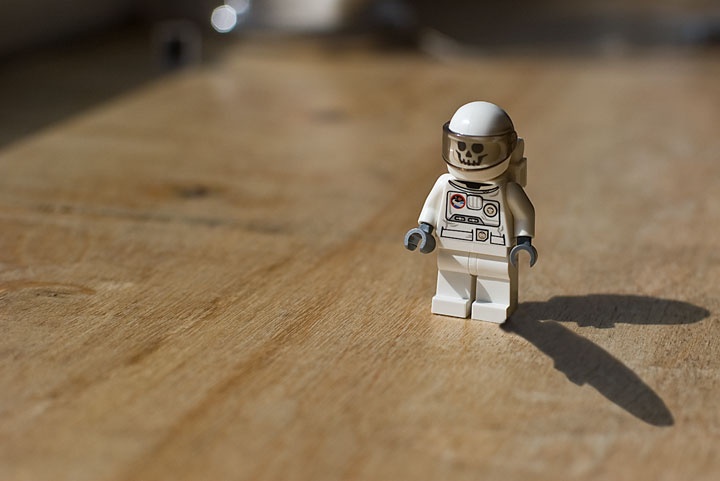A white LEGO astronaut minifigure with a skull face printed on the helmet stands alone on a wooden surface. The figure is in…