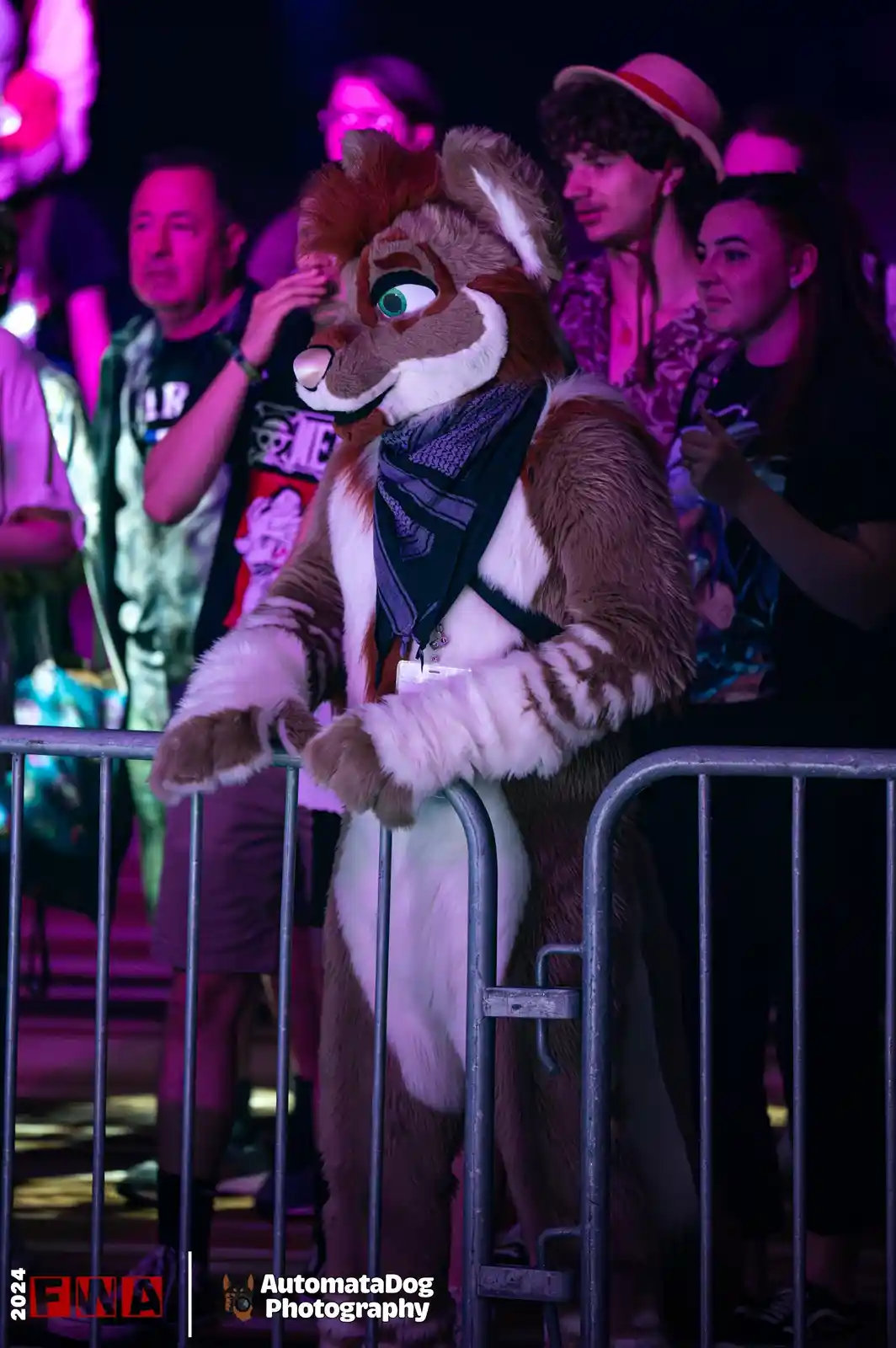 A person in a detailed brown and white fursuit with green eyes and a scarf leans on a metal barrier at a crowded event under…
