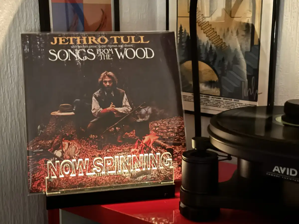 A vinyl record album cover for Jethro Tull's 'Songs from the Wood' is displayed upright on a red shelf next to a black AVID…