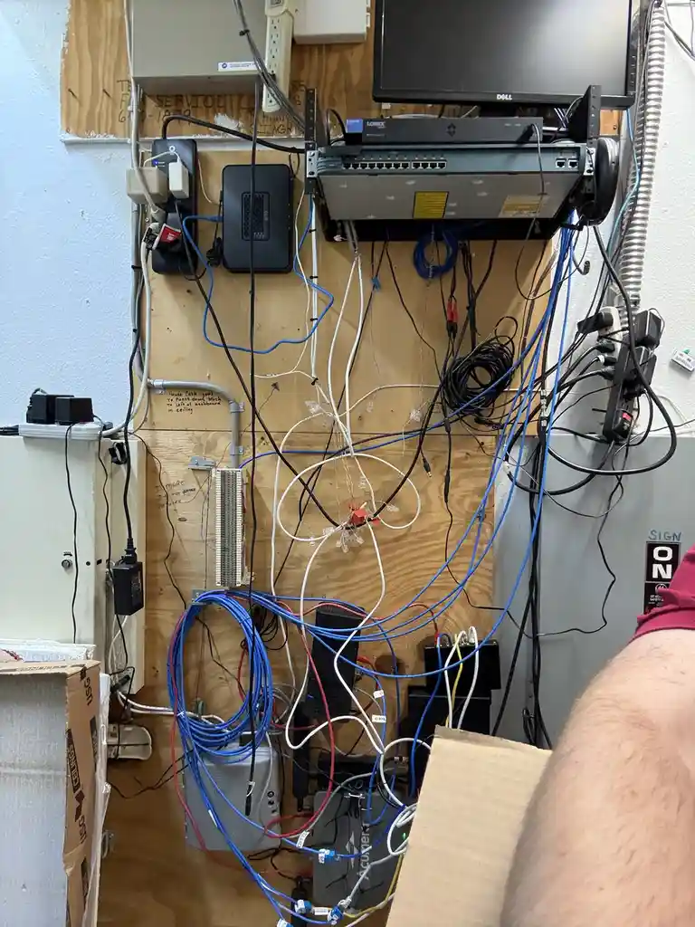 A chaotic wall of tangled blue, black, white, and red network cables connected to various routers, switches, and power…