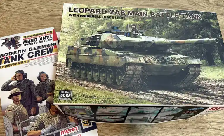 Two model kit boxes are shown on a wooden surface: one for a Leopard 2A6 main battle tank and another for a modern German…