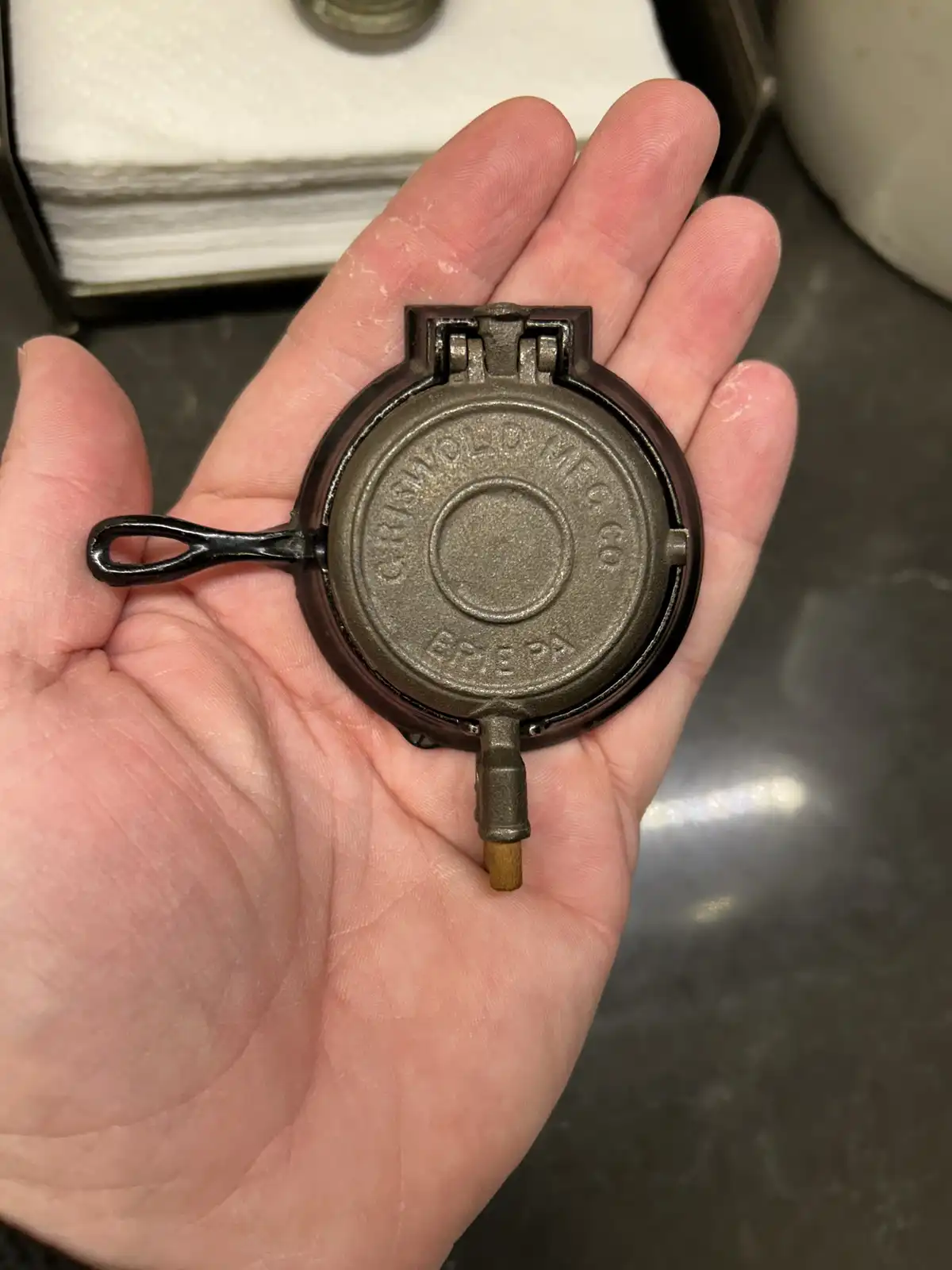 A person holds a small, round, cast-iron Griswold waffle iron in their open palm, showing its engraved brand and model. The…