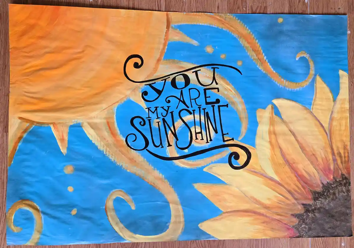 A hand-painted artwork on paper depicts a large, stylized sun with orange-yellow rays against a blue sky with small yellow…