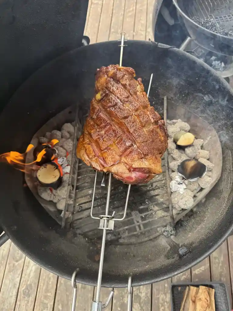 A large, scored piece of meat is roasting on a metal spit over glowing charcoal in a black kettle grill, with flames and…