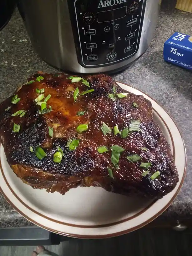 A large, glazed, charred brisket sits on a white plate with brown trim, garnished with chopped green onions. In the…