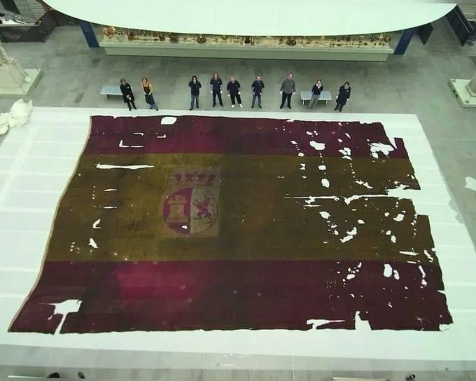 an overhead view of a large, tattered, and faded historical flag of spain displayed on a white platform in a museum. Several…