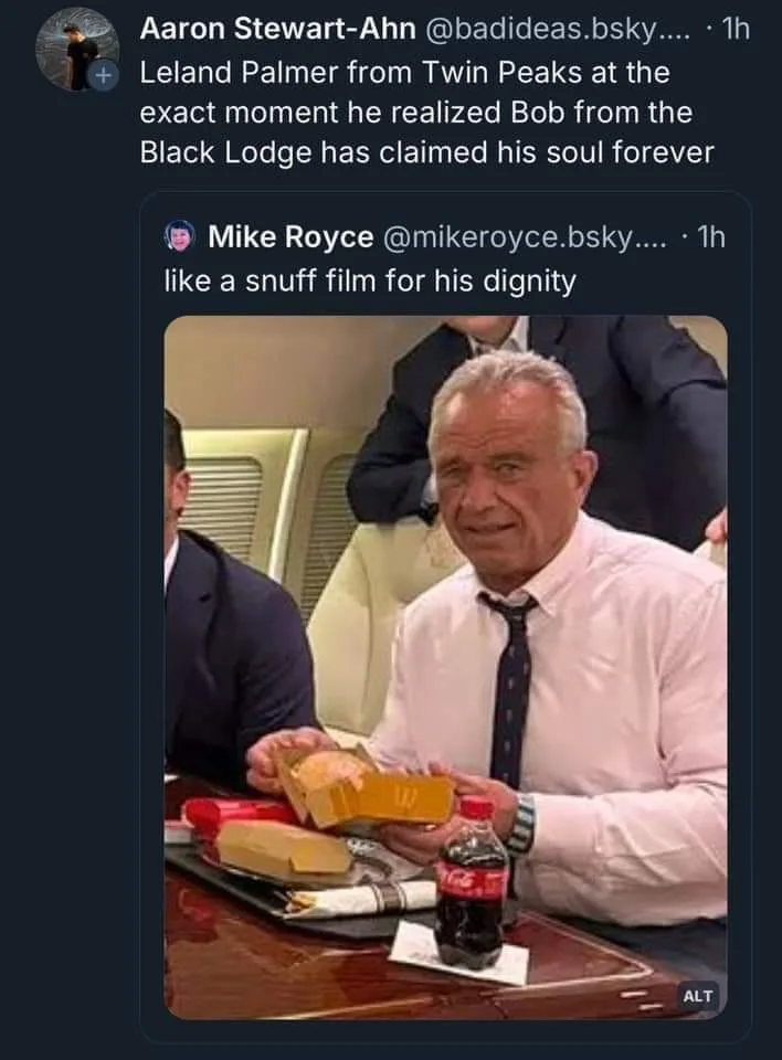 An older man with gray hair, wearing a white shirt and dark tie, sits at a table holding a partially eaten sandwich in a…