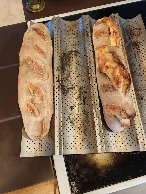 Two baguettes sit on a perforated metal baking tray, one on the left is evenly golden-brown, while the one on the right is…