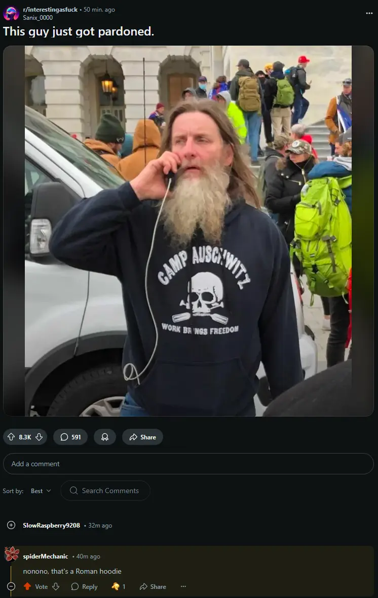 A man with a long beard and dark hoodie stands outdoors near a white vehicle, holding a phone to his ear. His hoodie…