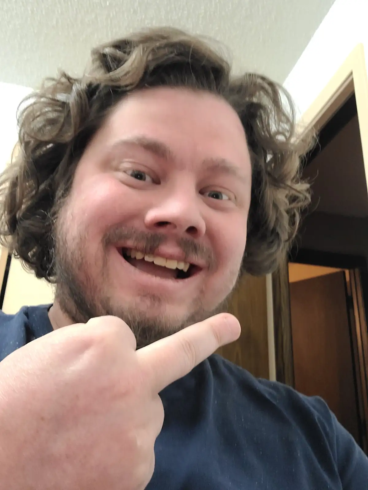 A man with curly brown hair and a beard smiles widely at the camera while pointing to his own mouth with his index finger…