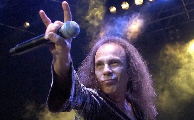 a man with long curly hair and a shiny jacket holds a microphone and makes a rock hand gesture while looking at the camera…