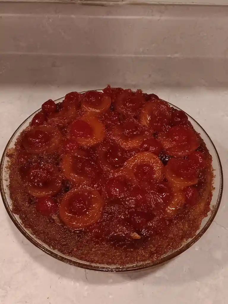 A round, glass dish holds a baked dessert topped with glossy red cherries and orange apricot slices, sitting on a white…