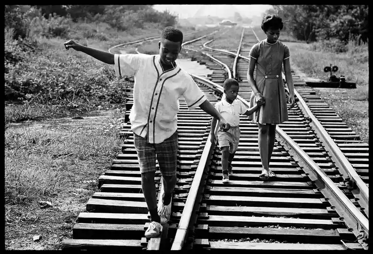 A black-and-white photograph shows three Black children walking along railroad tracks. An older boy in a baseball jersey…