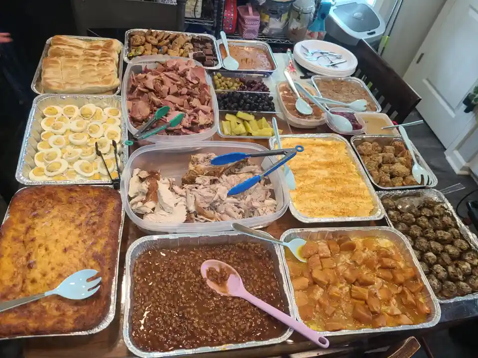 A table is covered with numerous aluminum trays filled with various foods including deviled eggs, sliced ham, pulled pork…