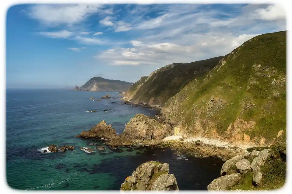 A scenic coastal landscape with green hills, rocky cliffs, and blue ocean under a partly cloudy sky. The image has a white…