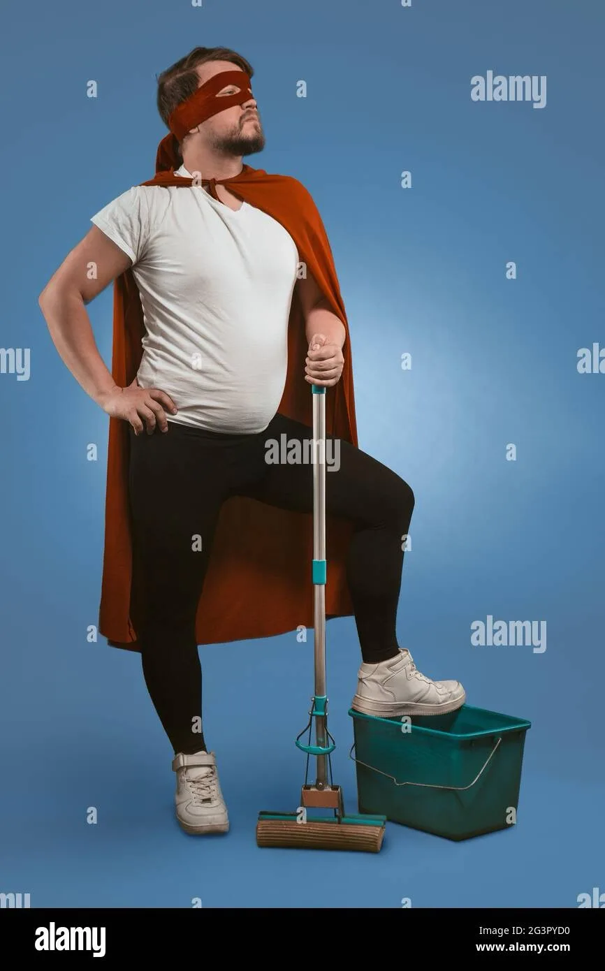 A man dressed as a superhero with a red cape and blindfold stands confidently with one foot on a green mop bucket, holding a…