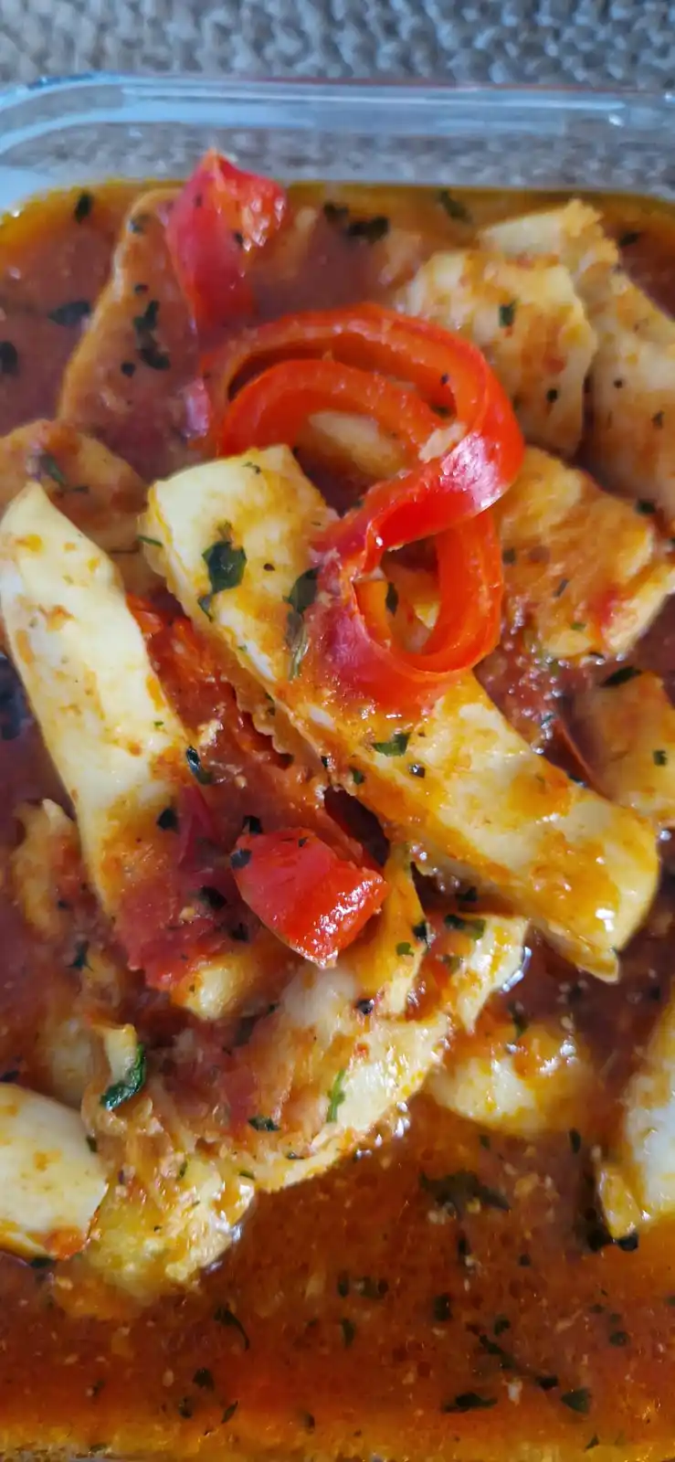 A close-up photo of a glass dish containing cooked white fish fillets in a red tomato-based sauce, garnished with sliced red…