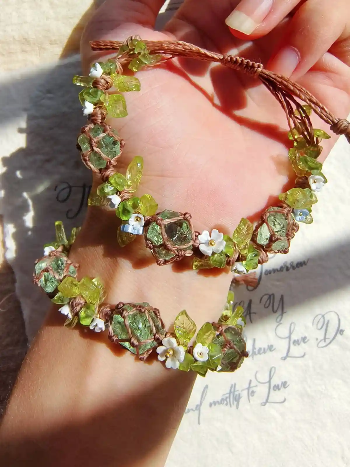 A hand holds a handmade macrame bracelet featuring green gemstones, white flower beads, and brown cord, set against a…