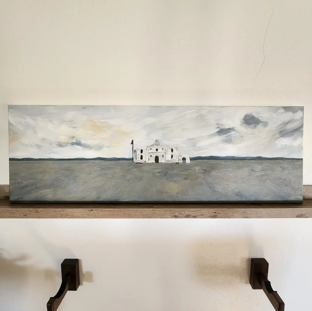 A long, horizontal painting of a lone, white, dilapidated building in a vast, gray, empty landscape hangs on a white wall…