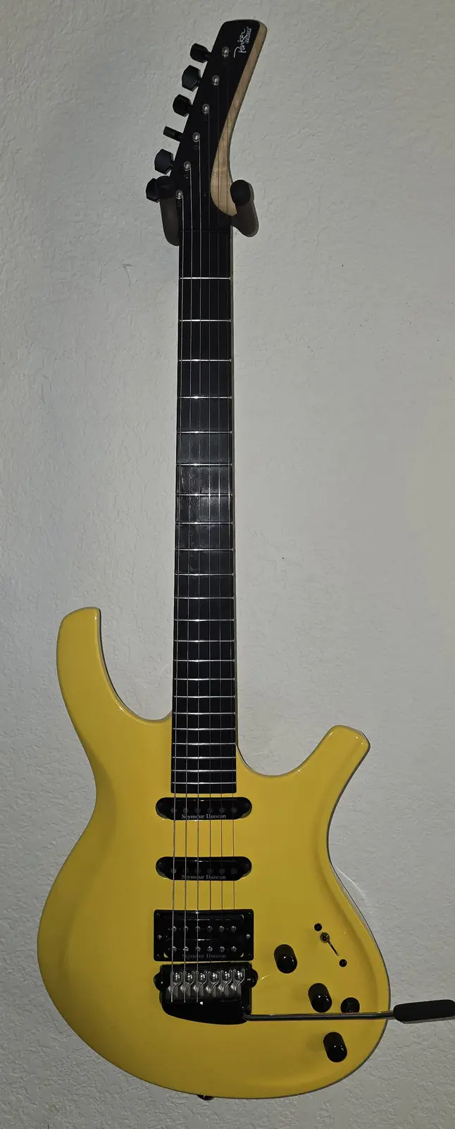 A bright yellow Parker guitar with black hardware and a black fretboard is mounted vertically against a textured white wall…