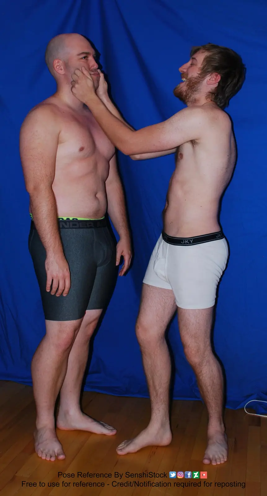 Two shirtless men stand side-by-side against a blue backdrop, one with a beard laughing while holding the other's face. The…