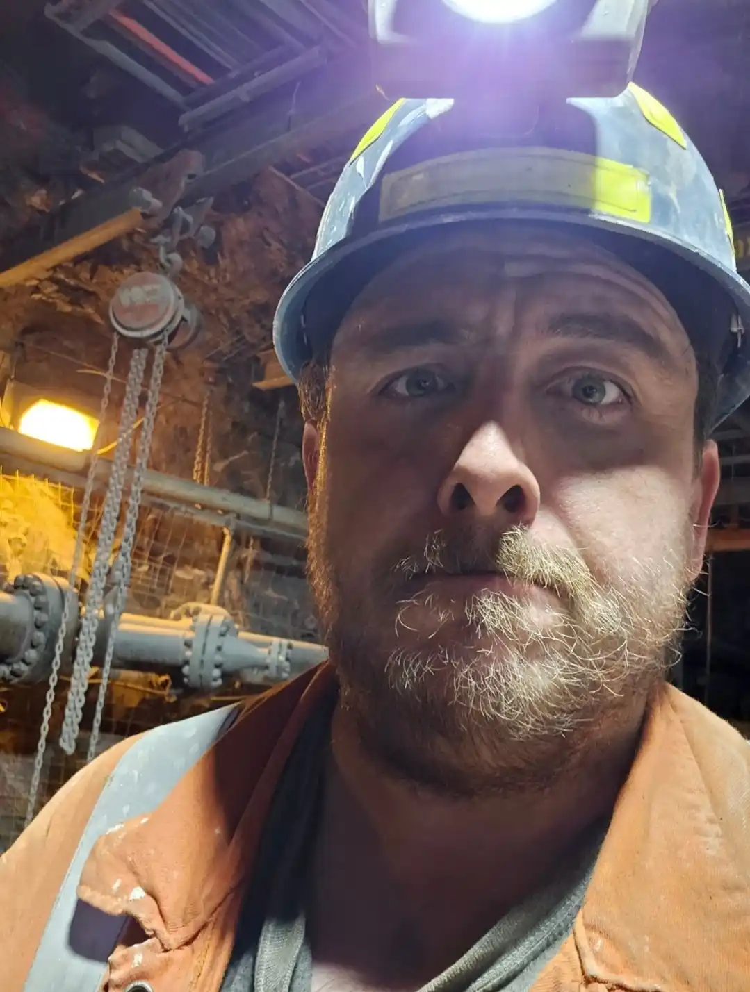 A close-up selfie of a man wearing a blue hard hat with a headlamp and an orange work vest, set against the backdrop of a…