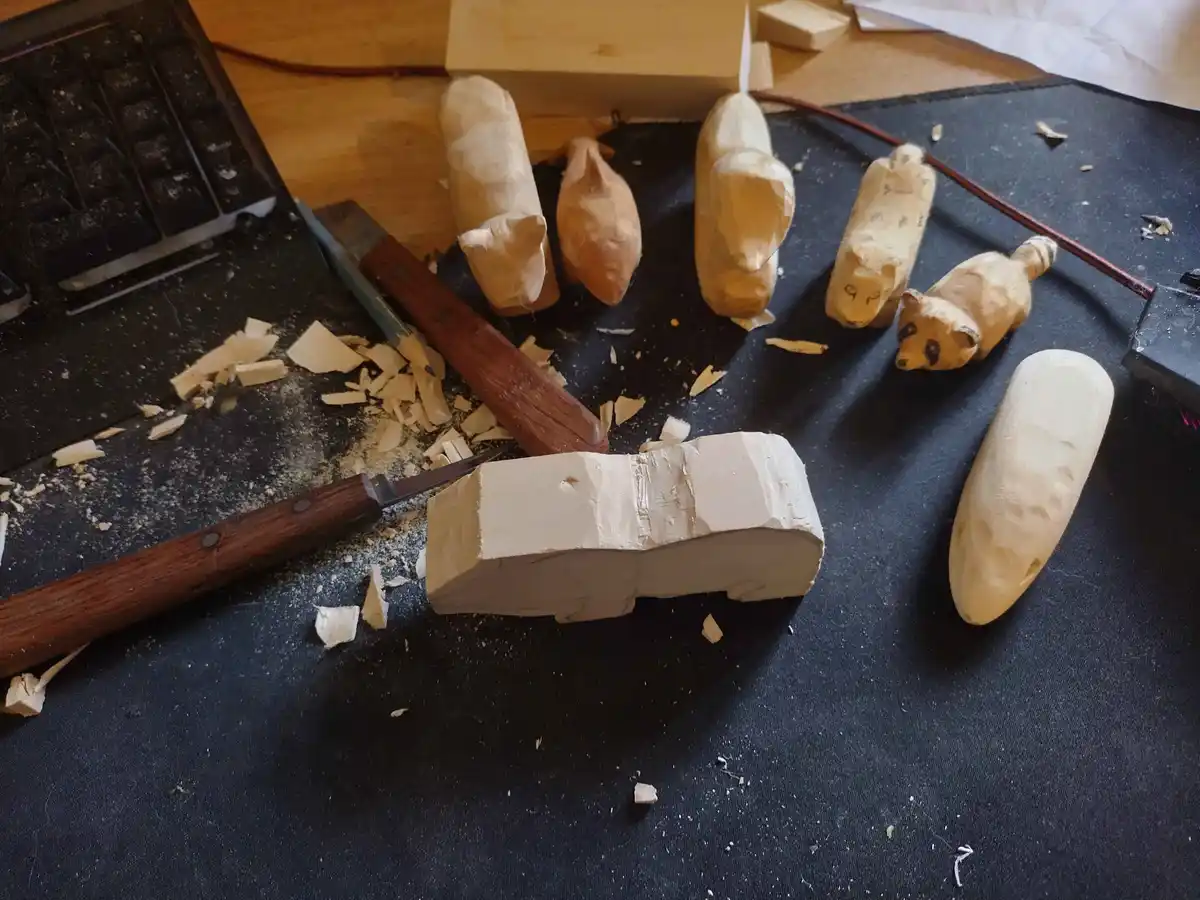 A wooden carving project shows a block of wood shaped like a car in the foreground, surrounded by various other carved…
