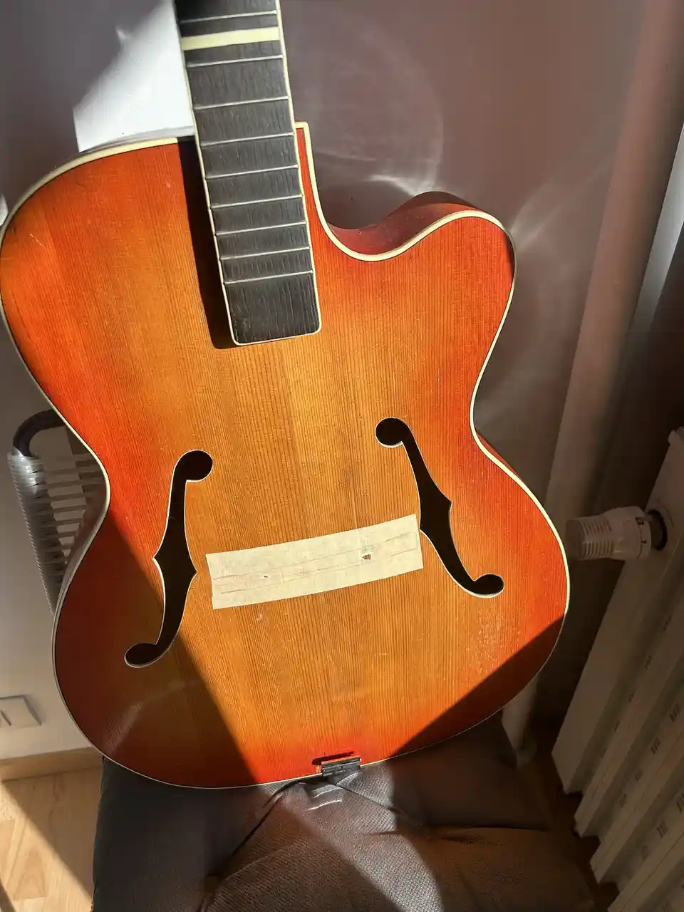 A close-up photo of an orange sunburst archtop guitar with f-holes, resting on a chair, has a piece of tape covering a small…