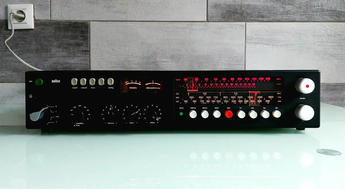 A vintage Braun radio receiver with a black panel, numerous knobs, and glowing red frequency dials sits on a reflective…