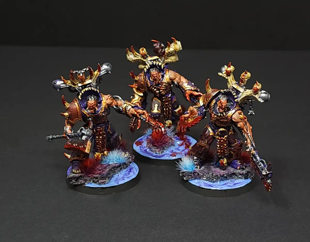 Three intricately painted fantasy miniatures, resembling demonic or warlike figures with ornate armor and weapons, stand on…