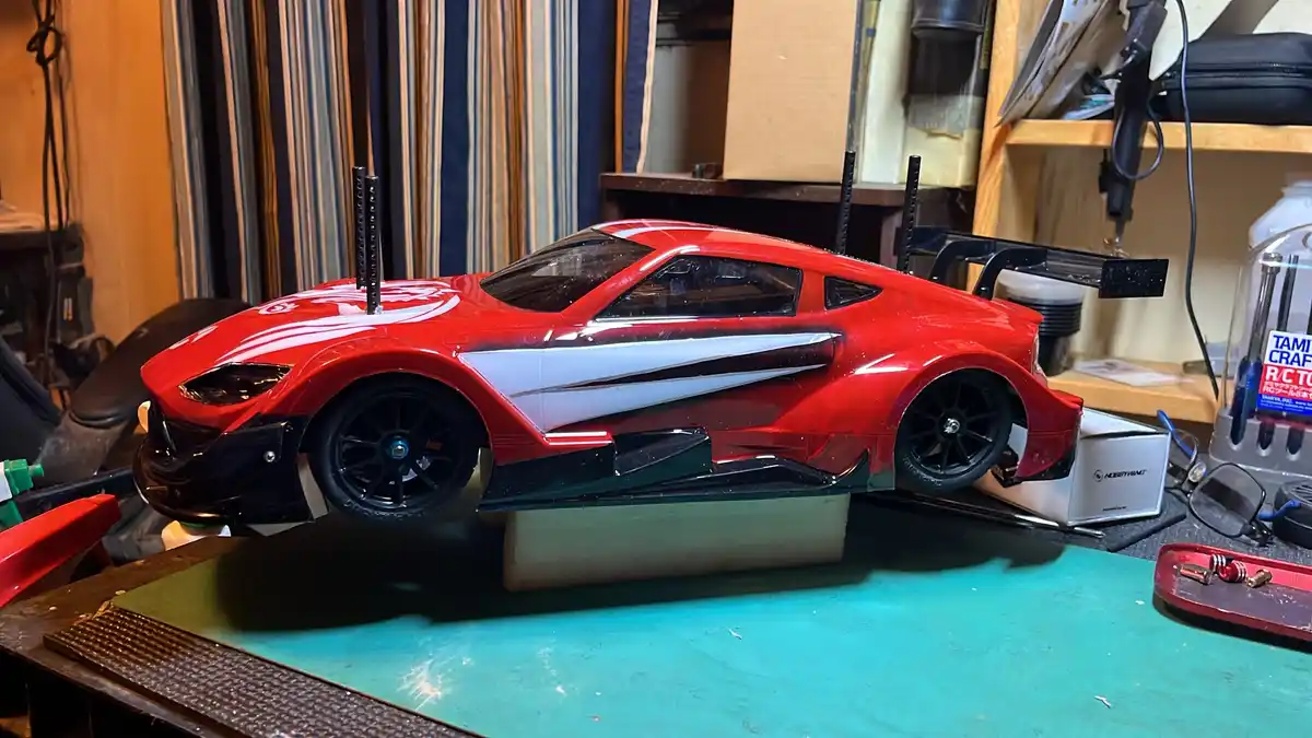 A red and white radio-controlled car with black wheels and a large rear wing sits on a workbench in a cluttered workshop…
