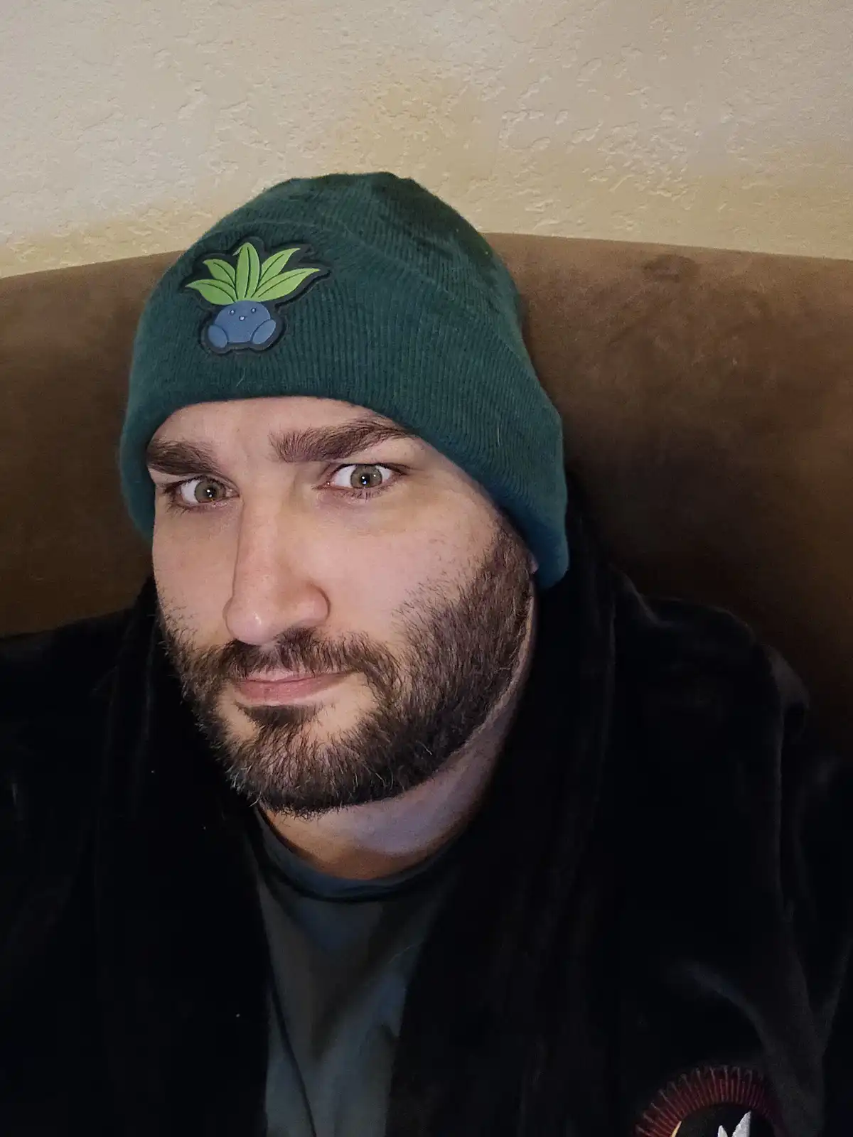 A man with a beard and intense stare wears a green beanie featuring the Pokémon Bulbasaur logo, sitting on a brown couch…