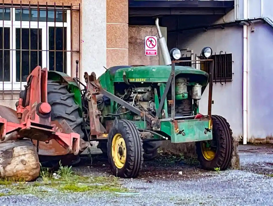A green John Deere tractor is parked outdoors next to a building, with its front end facing the viewer and its rear end…