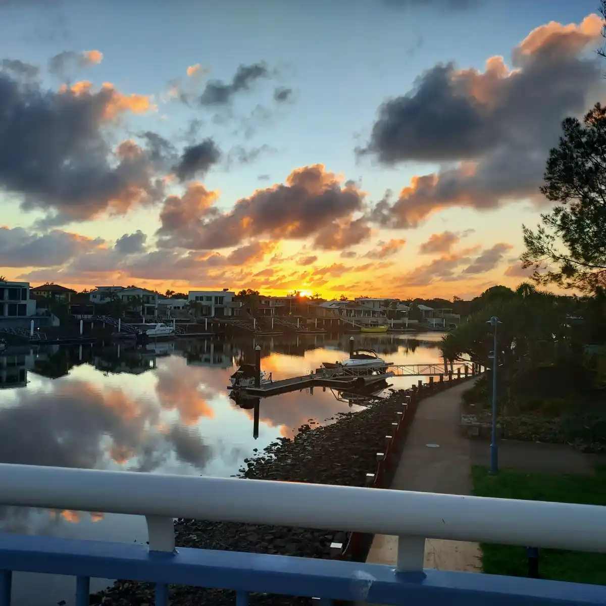 A serene sunset over a waterfront with houses, docks, and boats, their reflections shimmering in calm water. The sky is…