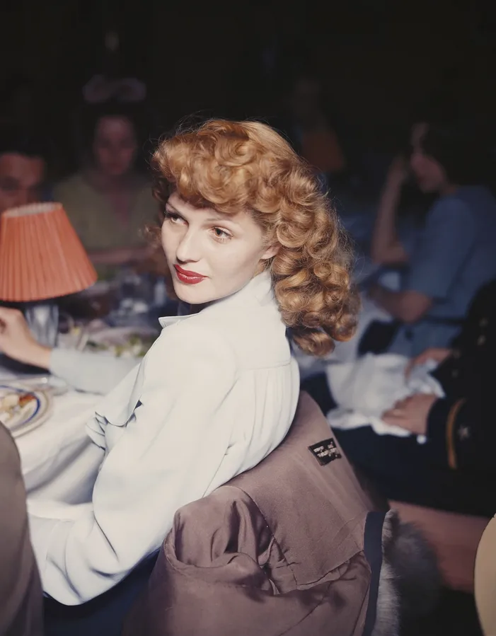 A woman with curly blonde hair and red lipstick looks over her shoulder at the camera while seated at a dinner table. She is…