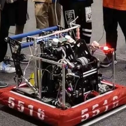 A complex, homemade robot with exposed wires, motors, and a red LED light is mounted on a red inflatable platform marked…