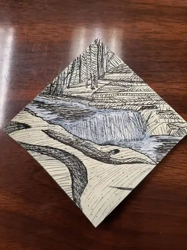 A hand-drawn sketch on a diamond-shaped piece of paper, rotated 45 degrees, depicts a waterfall cascading over rocks with…
