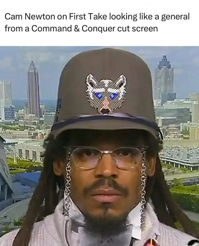 A man with dreadlocks and glasses wears a brown military-style hat with a pixelated raccoon face emblem and chains hanging…