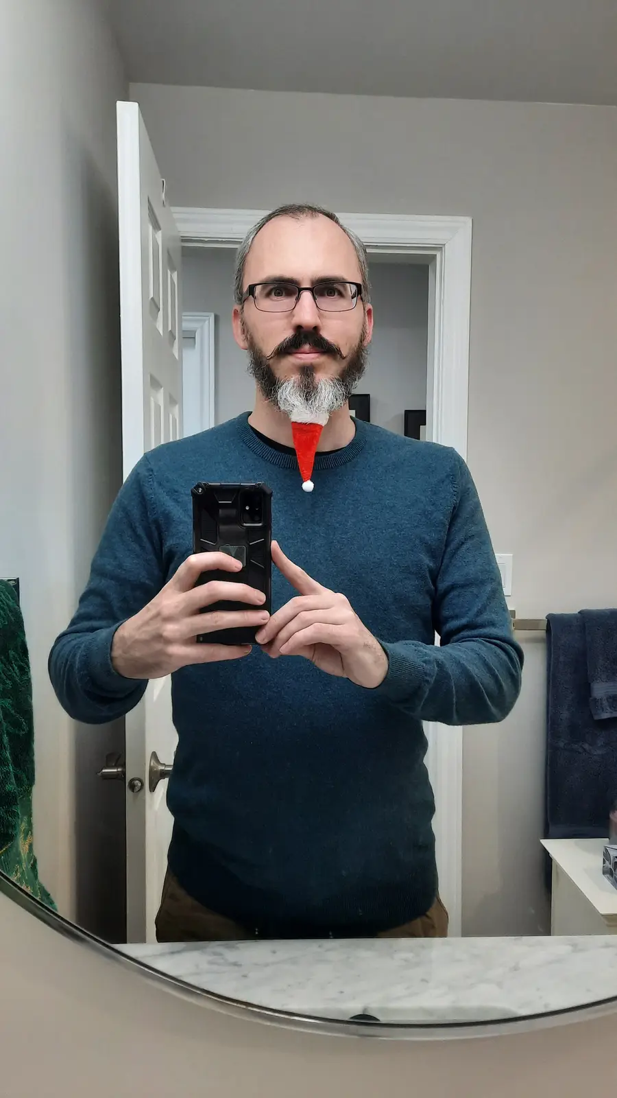 A man with glasses and a goatee takes a mirror selfie in a bathroom, holding a phone. A small red Santa hat is stuck to the…